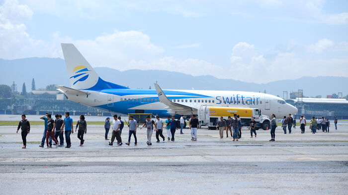 Groups of asylum seekers walk across an airport tarmac near a Swiftair plane, illustrating immigration playbook and global bargaining chips.