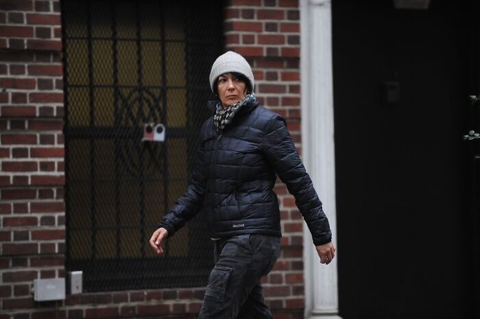 Ghislaine Maxwell wearing a black jacket and gray beanie, walking past a brick building in an urban setting.