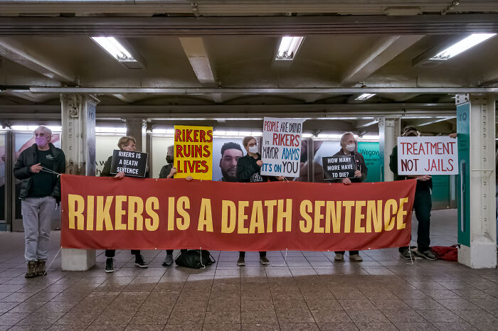 Protesters holding signs highlighting systemic dysfunction at Rikers Island, including calls for treatment instead of jail.