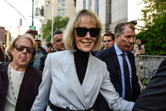 Woman in light gray blazer and black sunglasses walking outdoors, surrounded by people, related to Trump accusations and Epstein context.