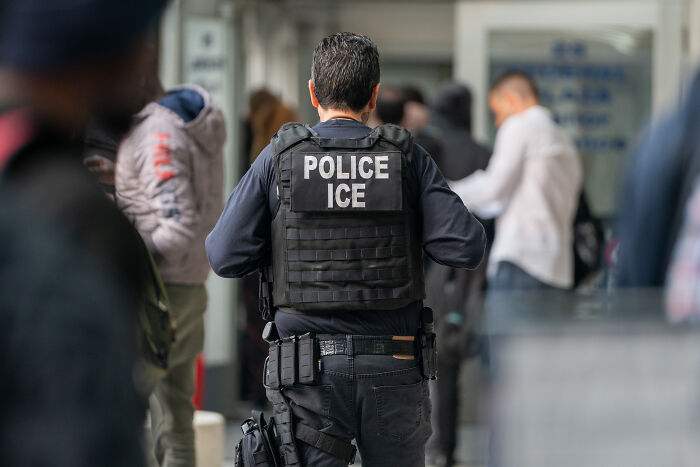 ICE police officer in tactical gear walking through a crowded area related to immigration sanctuary law enforcement efforts.