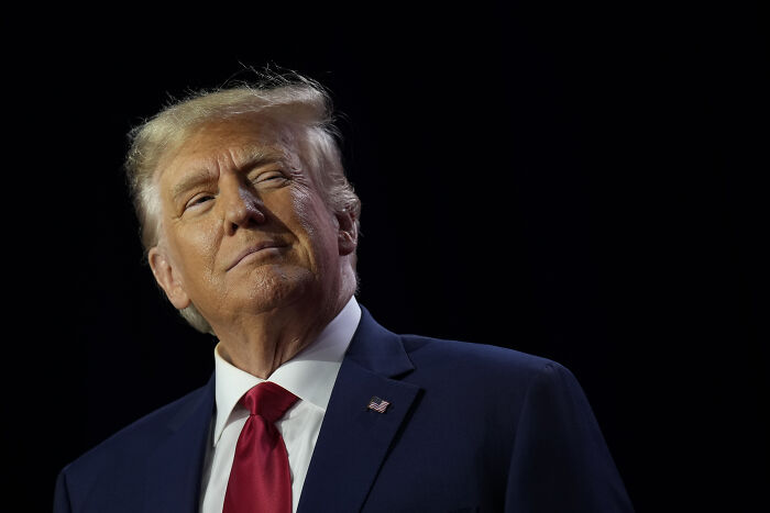 Donald Trump in a dark setting, wearing a blue suit and red tie, highlighting old accusations amid Epstein context.