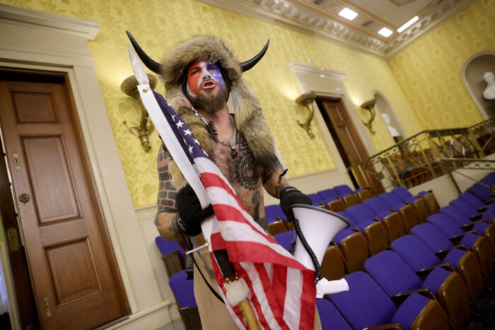 Man dressed as QAnon Shaman with horned fur hat and face paint holding American flag inside a room with blue chairs.