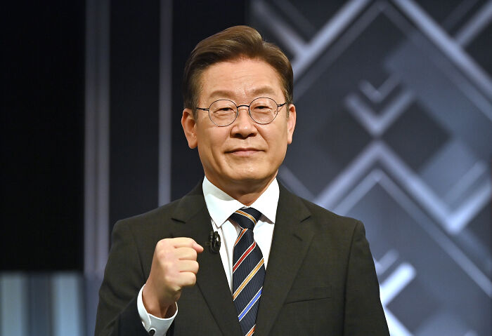 South Korean official in a dark suit and striped tie, making a fist gesture during a formal event or speech.