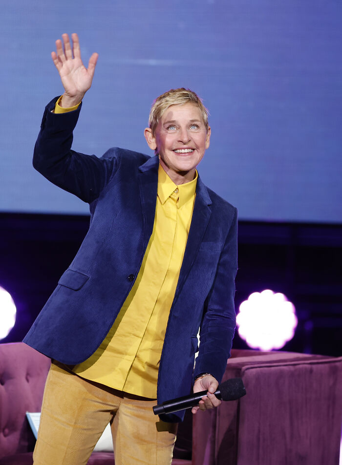 Ellen DeGeneres waving on stage in a blue jacket and yellow shirt during a live event with microphone in hand.