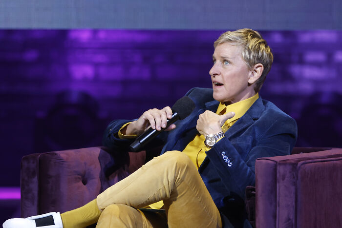 Ellen speaking into a microphone on stage, wearing a blue jacket and yellow shirt, engaging with the audience.