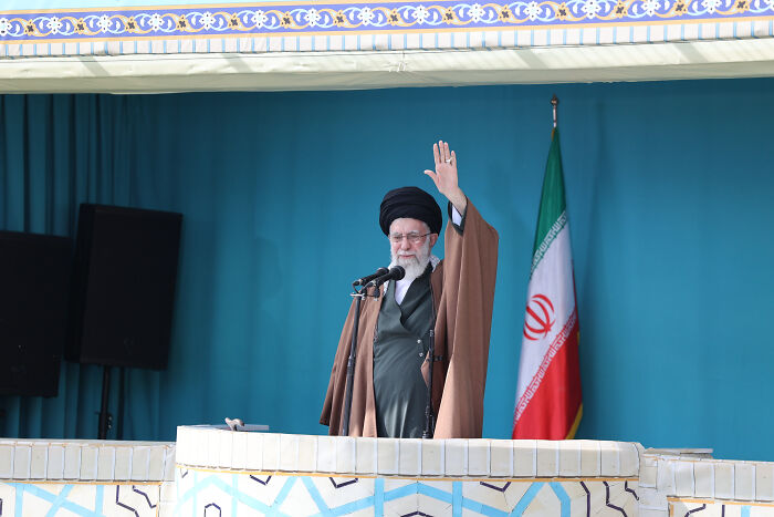 Religious leader speaking at podium with Iran flag in background, amid Trump world hacked staff emails threat news.