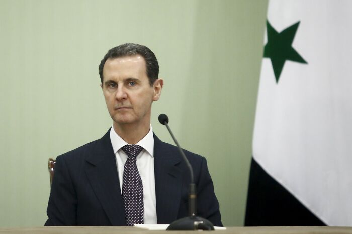Syrian leader Bashar al-Assad seated near a microphone with Syrian flag, amid ongoing fighting and Israel involvement in Syria.