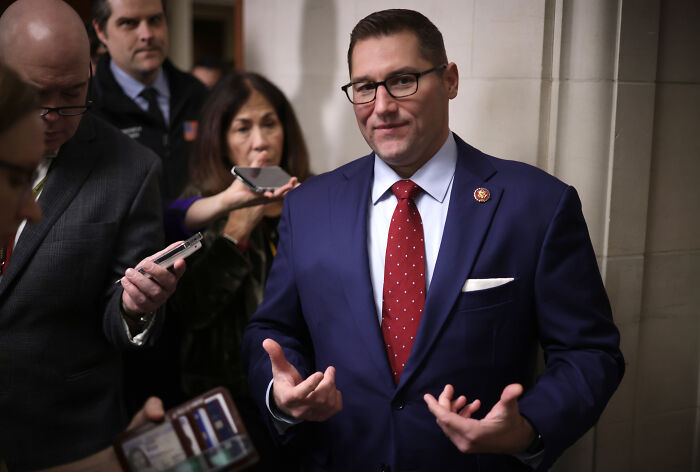 Republican lawmaker speaking to reporters in a hallway, wearing glasses and a navy suit with a red tie.