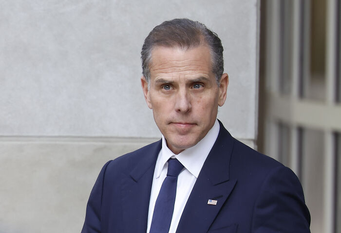 Hunter Biden in a navy suit and tie, standing outdoors near a building, looking serious and focused.