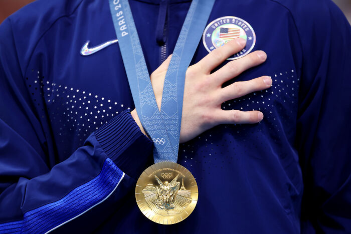 Athlete wearing U.S. Olympic jacket holding gold medal and touching badge, representing U.S. Olympic Committee sports policy.