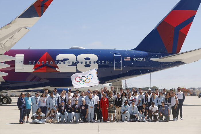 Group of people holding Olympic flag in front of L28 plane symbolizing U.S. Olympic Committee and transgender athlete ban in women&rsquo;s sports.