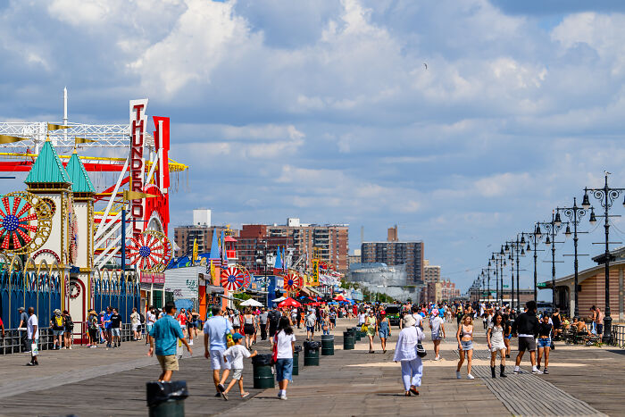 Crowded Coney Island boardwalk with families and visitors amid a sunny day, near amusement rides and shops.