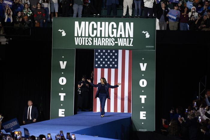 Kamala Harris on stage at Michigan event with American flag backdrop during political campaign vote rally.