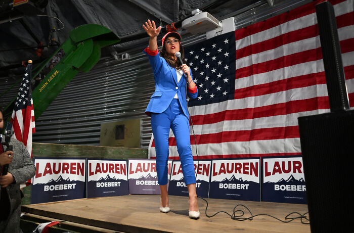 Congresswoman Lauren Boebert speaking on stage with American flag and campaign signs in the background.