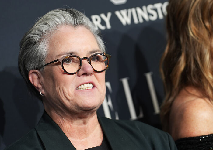 Ellen wearing glasses and a dark blazer, speaking at an event, stunning fans by siding with Rosie O&rsquo;Donnell.