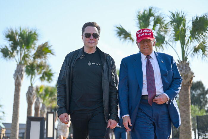 Elon Musk wearing sunglasses and black jacket walking outdoors alongside man in suit and red MAGA hat