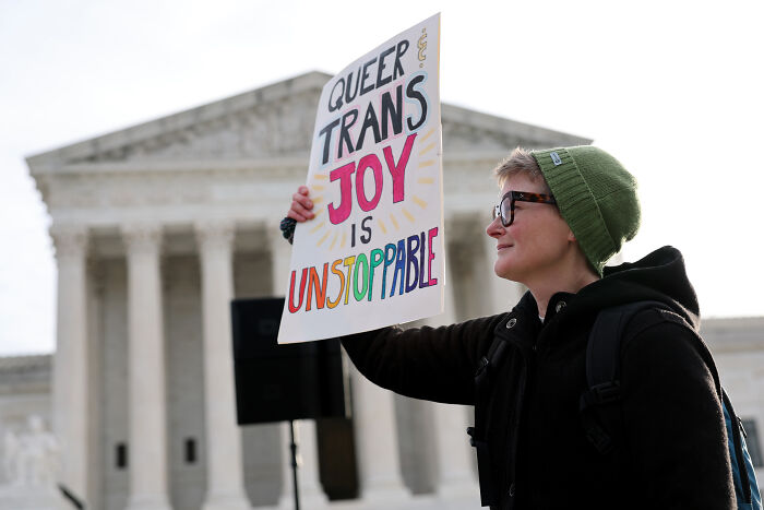 Person holding a sign about trans joy protesting outside a government building amid DOJ subpoenas for doctors providing care to trans minors