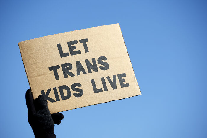 Hand holding a cardboard sign with Let Trans Kids Live text against a clear blue sky related to doctors providing health care to trans minors
