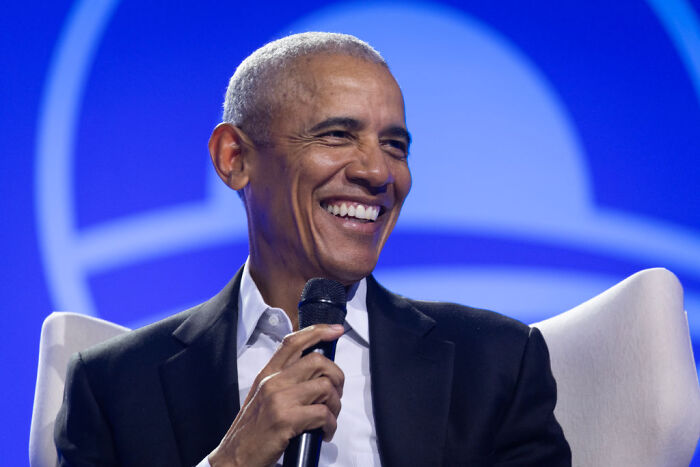 Barack Obama smiling and holding a microphone during a public speaking event with a blue background.