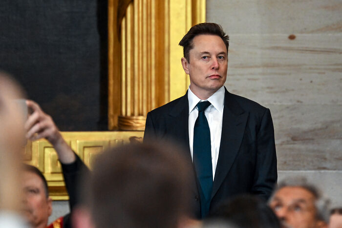 Elon Musk in a dark suit standing in a formal setting, related to X boss Linda Yaccarino's resignation news.