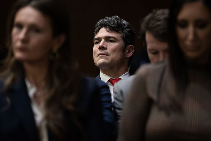 Man in a suit and red tie attends a formal hearing amid blurred individuals, linked to January 6 conspiracy theorist appointment.
