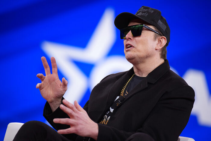 Elon Musk wearing sunglasses and a cap, speaking passionately with hand gestures against a blue star background.