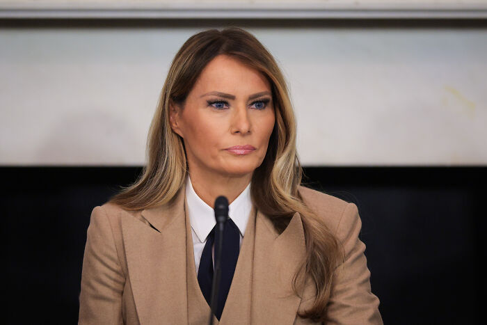 Melania Trump wearing tan blazer and white shirt, seated indoors with a serious expression on her face.