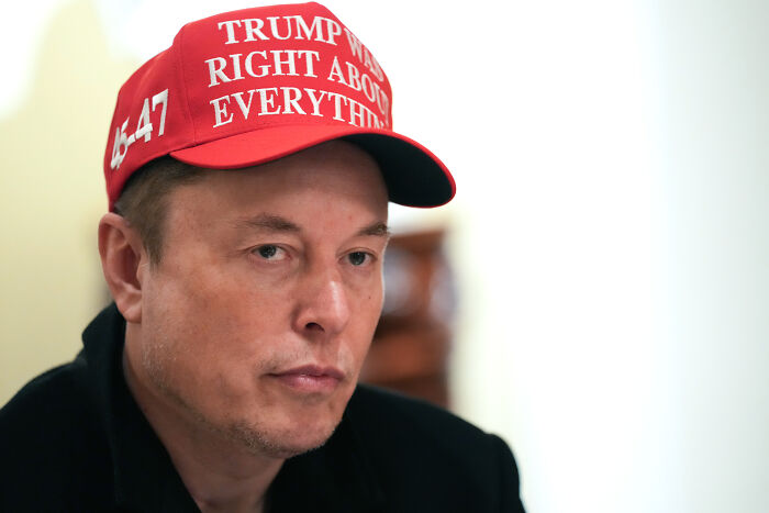 Elon Musk wearing a red cap with political text, looking serious amid discussions about the debt bill and America party.