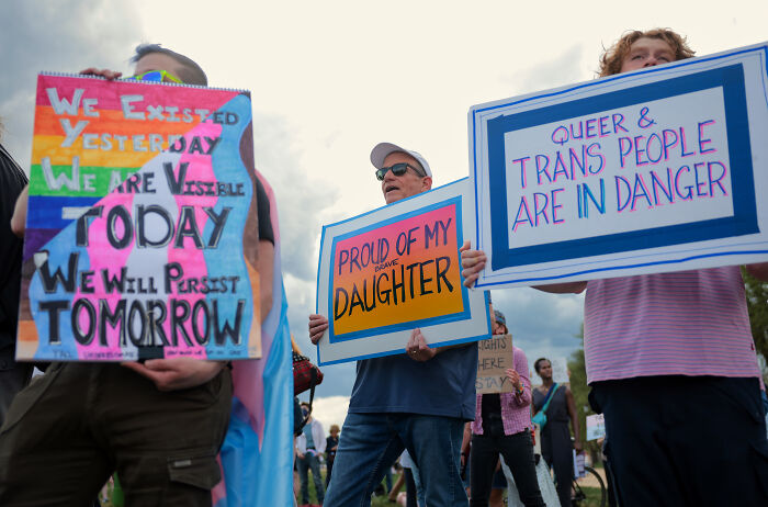 Protesters holding signs about trans rights, highlighting the impact of Trump's anti-trans agenda on lives.