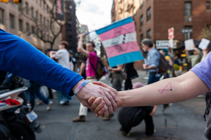 Two people holding hands in a city protest with a transgender pride flag, representing the impact of Trump&rsquo;s anti-trans agenda.