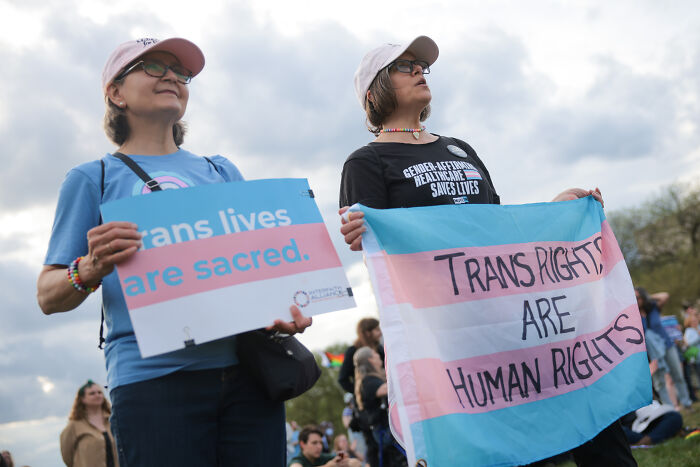 Two protesters holding signs advocating for trans rights and highlighting the impact of Trump&rsquo;s anti-trans agenda.