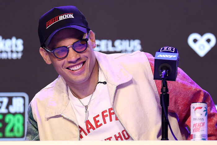 Boxing champion smiling at a press conference wearing a cap and glasses amid gun case and cartel link allegations.