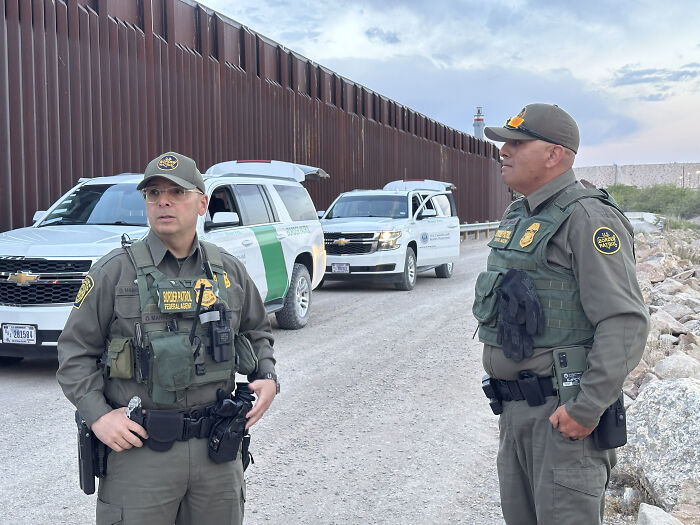 Two Border Patrol agents in uniform standing near U.S.-Mexico border fence with patrol vehicles, highlighting asylum enforcement issues.