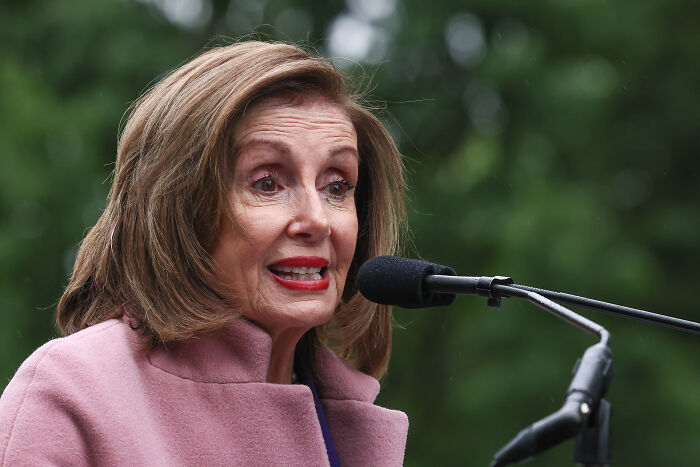 Speaker Nancy Pelosi in a pink coat speaking at a microphone on congressional stock trading ban discussion outdoors.
