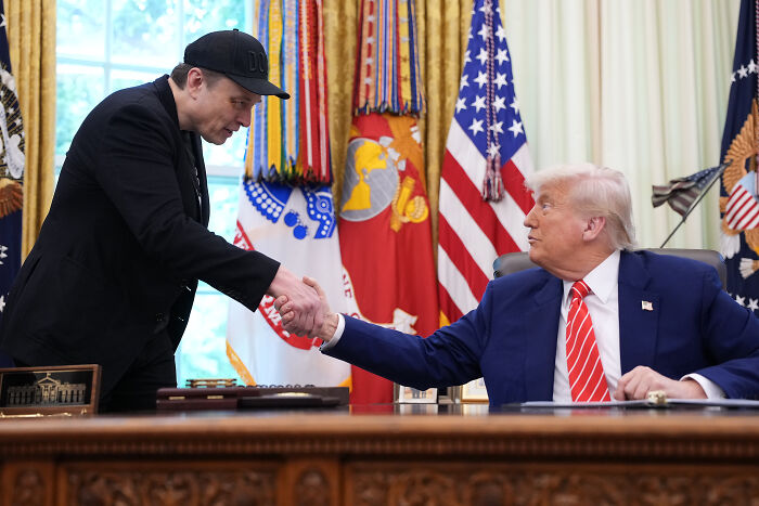 Elon Musk and Donald Trump shaking hands in the Oval Office during a meeting about DOGE and billionaires.