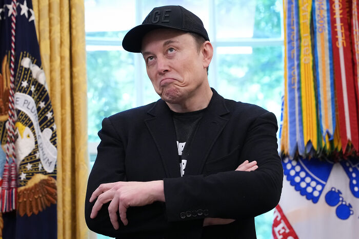 Elon Musk wearing a black cap and jacket, standing with arms crossed in a room with flags and a window in the background.