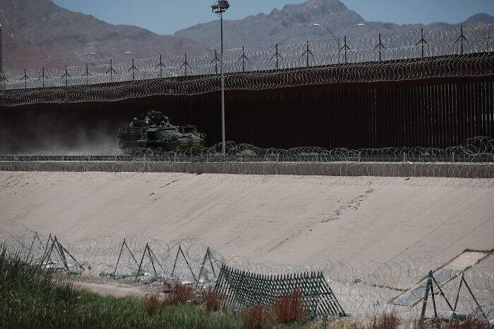 Armored vehicle driving along a barbed wire fence securing the U.S.-Mexico border with mountains in the background.
