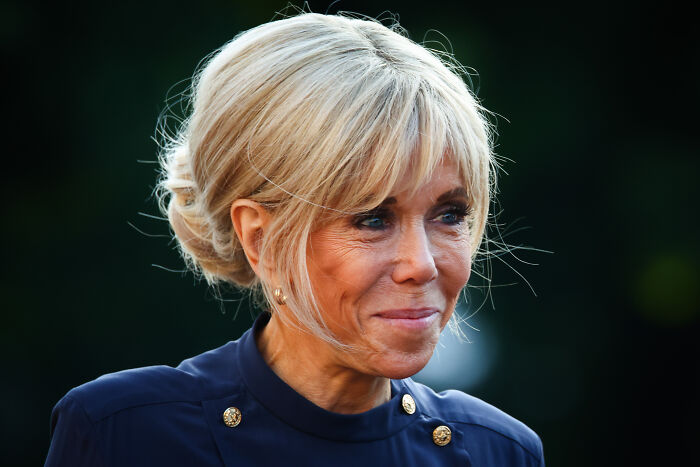 Brigitte Macron wearing a navy outfit and smiling softly in an outdoor setting with a dark blurred background