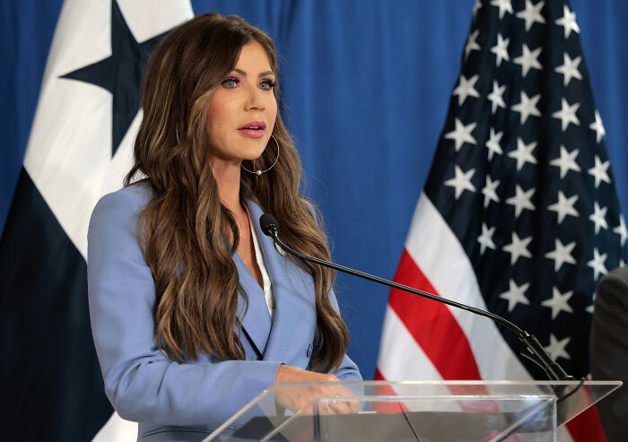 Kristi Noem speaking at a podium with Texas and American flags behind her during a press event about flood aid delay.