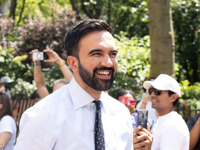NYC mayoral nominee smiling outdoors with crowd in background amid Trump calling him a communist controversy.