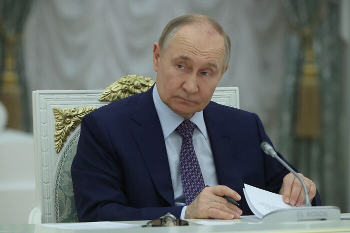 Vladimir Putin in a formal setting, wearing a dark suit, with papers and a microphone, related to Moscow threat audio.