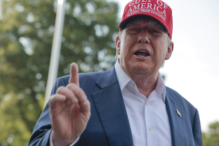 Donald Trump wearing a red Trump hat and blue suit, speaking outdoors with hand raised, referencing NYC mayoral nominee and communism.