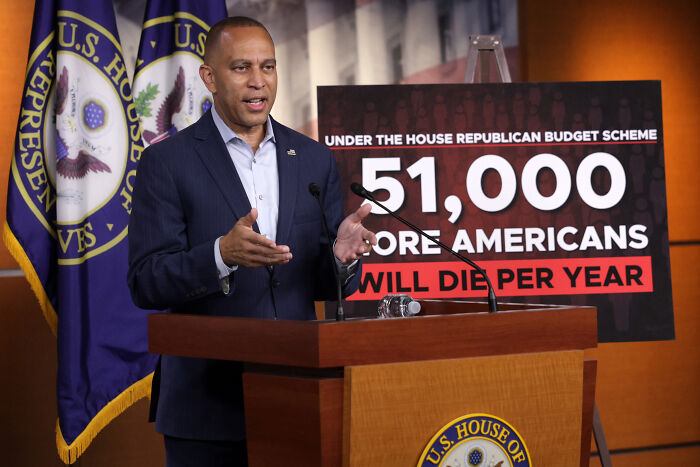 Congress member speaking at podium with U.S. House flags and sign showing impact of House Republican budget scheme.