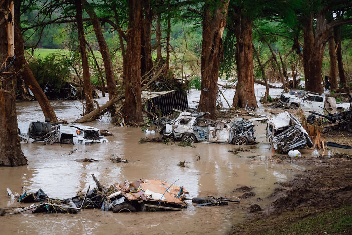 Flooded area with destroyed vehicles submerged in muddy water surrounded by trees, illustrating drowning flood victim controversy.