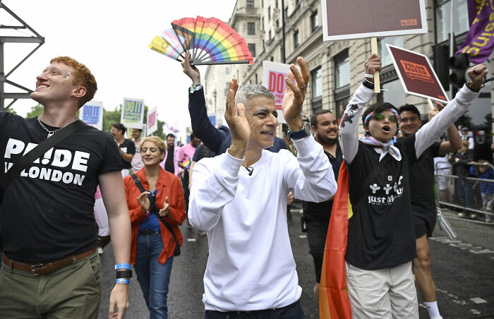 London Mayor at public event, surrounded by supporters holding signs and rainbow flags, celebrating with applause and cheers
