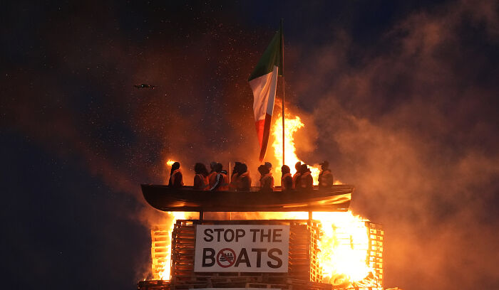 Bonfire with mock migrant boat and Stop the Boats sign set alight, sparking outrage over racist display at night.