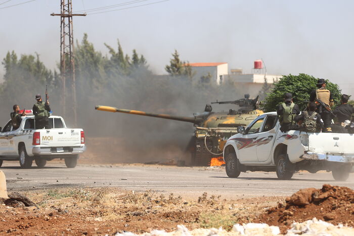 Armed fighters in pickup trucks near a tank firing in a dusty battlefield during ongoing fighting in Syria.