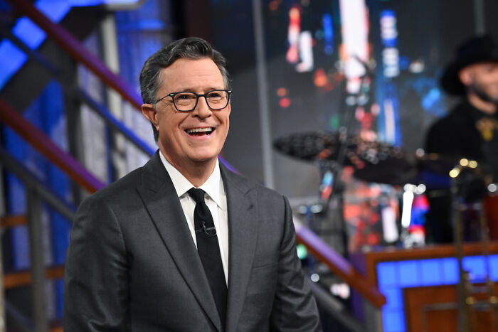 Stephen Colbert smiling on a TV set, dressed in a suit and glasses, related to Trump&rsquo;s quest to sue on TV.