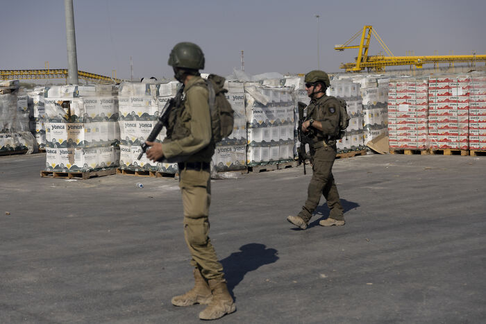Two armed soldiers in military gear standing near stacked supplies, highlighting Gaza situation and genocide issues.